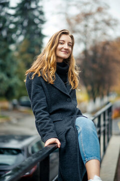 A woman in a long gray coat and blue jeans on white sneakers sits on the railing and looks at the camera on the street of an autumn city