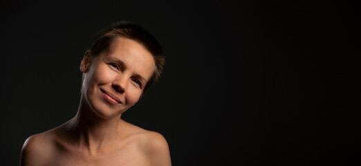 Capturing a middle-aged lady, short-haired, baring her shoulders, sporting a subtle grin, set against a black background with an available banner for text