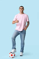 Handsome young man with soccer ball showing thumb-up gesture on blue background