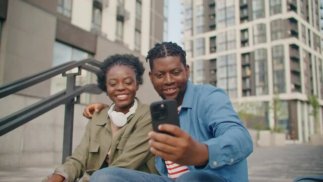 African American teenagers having fun on street. Sitting on staircase and making selfie with help of mobile device. Laughing while spending time together. Cute couple making photo.