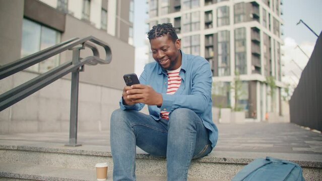 Zooming in on happy African American man sitting on stairs and typing message to someone. Smiling while using his cell phone. Spending time on fresh air with his technology device.