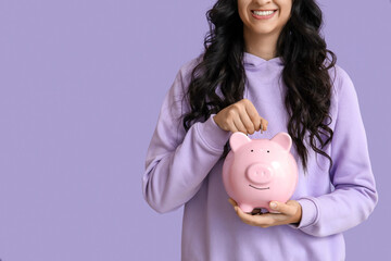 Pretty young woman putting coin into piggy bank on lilac background