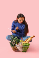 Young woman with shopping basket checking grocery list on pink background