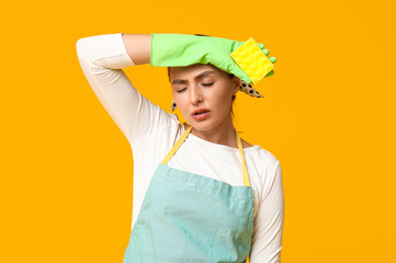 Portrait of tired young housewife in apron with rubber gloves and sponges on yellow background