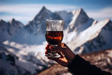 Foto op Plexiglas A hand holds a glass of mulled wine or hot black tea on snowy mountain peaks background. © OleksandrZastrozhnov