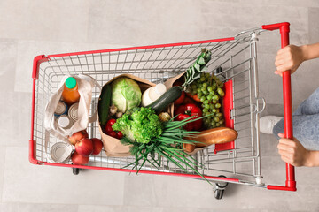 Young Asian woman with shopping cart and bags full of fresh food on tile floor, top view