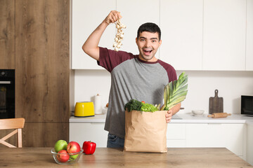 Happy young man with grocery bag holding fresh garlic at table in kitchen