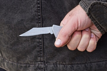 hand of a male criminal in black clothes and a gray knife behind his back on the street