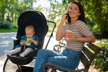 The baby is sitting in the stroller and holding a toy while mom is sitting on the bench and talking on the phone
