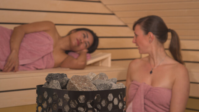 CLOSE UP, DOF: Hot heater stones and ladies wrapped in towels relaxing in sauna