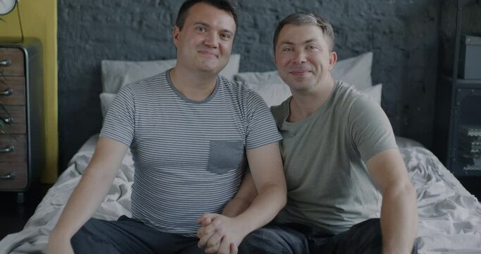Slow motion portrait of cute gay couple sitting in bed holding hands embracing and looking at camera. Love and LGBT relationship concept.