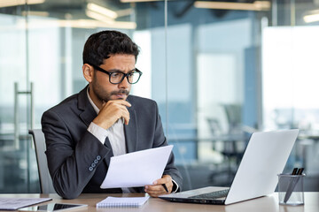 Portrait of serious pensive businessman behind paperwork, financier looking at documents, papers and contracts, thinking about solutions to set tasks, man inside office in business suit with laptop. - obrazy, fototapety, plakaty