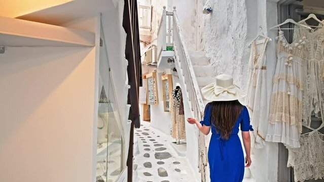 A tourist woman with a summer hat walking on the narrow streets with luxury shops in  Mykonos. Beautiful girl with a blue dress shopping for souvenirs, gifts and traditional Greek clothes in Mykonos.
