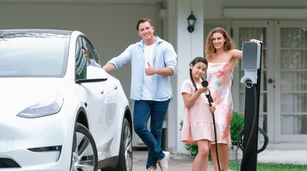 Modern family with environmental conscious recharging EV car with home EV charging station, Happy family with little young girl holding and pointing EV charger at camera. Synchronos - obrazy, fototapety, plakaty