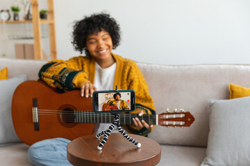 Blogger guitarist. African american girl blogger playing guitar talking to webcam recording vlog. Social media influencer woman streaming at home indoors. Music content creator broadcast tutorial - obrazy, fototapety, plakaty