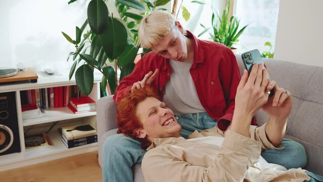 Smiling young gay couple with dyed hair taking selfies at cellphone on the sofa at home 