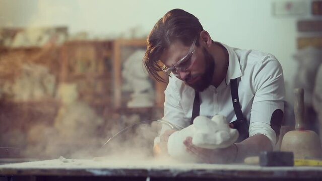 Pleasant attentive handsome modern bearded sculptor grinding stone sculpture in his creative workshop. Young man working with stone wearing protective glasses.