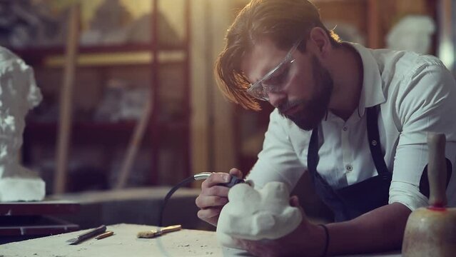 Pleasant attentive handsome modern bearded sculptor grinding stone sculpture in his creative workshop. Young man working with stone wearing protective glasses.