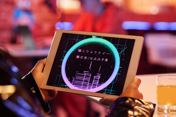 Screen of tablet with group of hieroglyphs in hands of young woman sitting in front of camera in cyberpunk bar or cafe at leisure