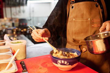 Close-up of young chef in apron putting mussels and other seafood into bowl with hot boiled ramen before serving meal to clients of cafe