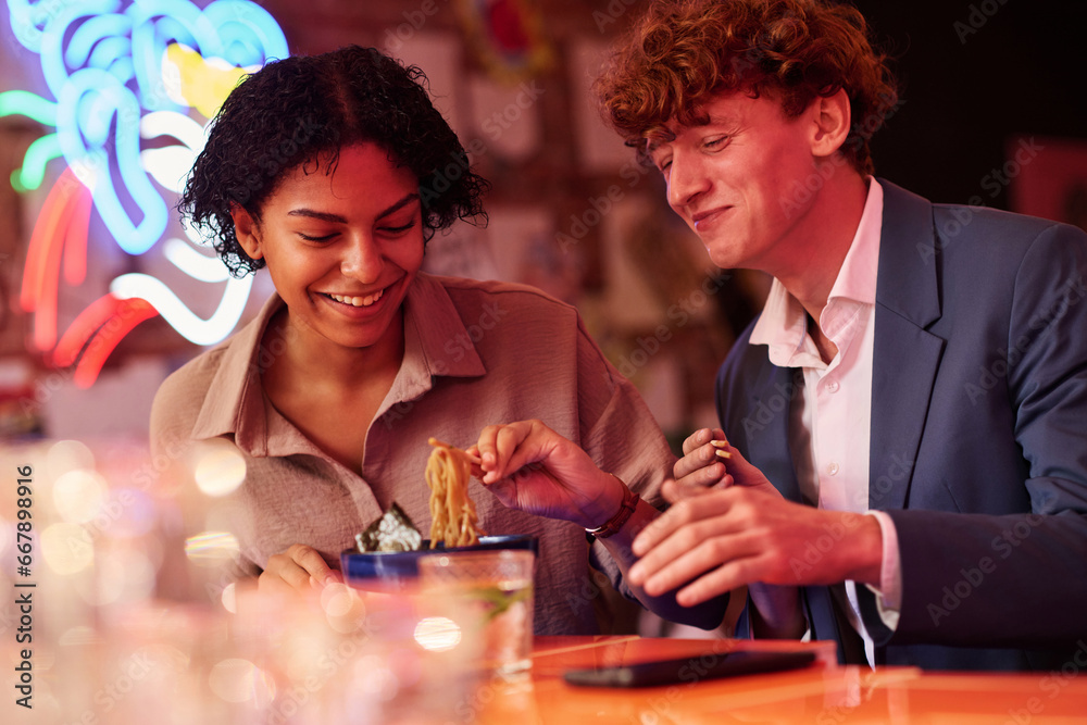 Poster happy young dates or colleagues sitting by table in japanese cafe or bar and having appetizing ramen
