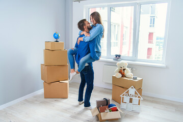 Front view of pretty smiling girl which holds by her handsome young boyfriend in the middle of their new flat full of cardboard boxes.