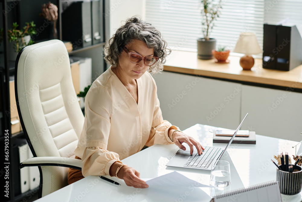 Wall mural mature female employer looking through paper document and typing on laptop keyboard while entering p