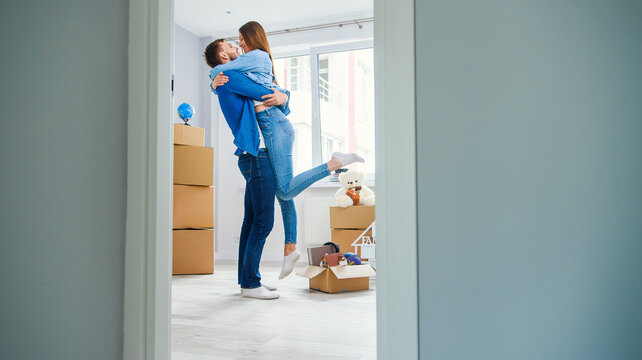 Front view of pretty smiling girl which holds by her handsome young boyfriend in the middle of their new flat full of cardboard boxes.