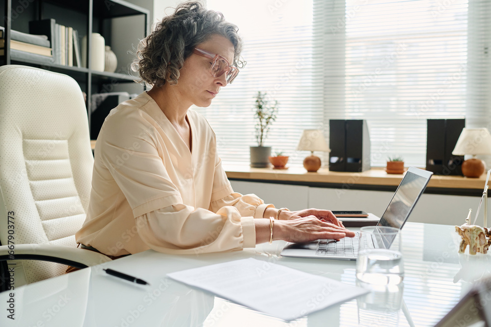 Poster serious mature female entrepreneur typing on laptop keyboard by workplace while entering data or mak