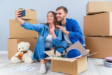 Fototapeta premium Young couple taking selfies in their new home while sitting among cardboard boxes. Happy attractive young couple is moving, making a selfie, cuddling and smiling.