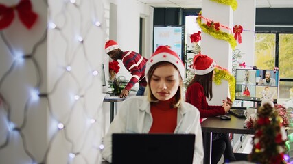 Asian staff member in videocall with teammates during Christmas season in festive decorated office. Employee in teleconference meeting with team in busy xmas ornate workplace - obrazy, fototapety, plakaty