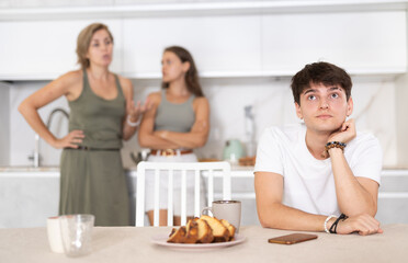 Obraz na płótnie Canvas Disgruntled and bored young man is sitting near table and has turned his back on his mother and sister. Young man suggests that arguing women calm down and find compromise