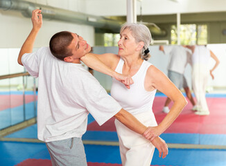 Focused elderly woman performing elbow strike and wristlock, painful control move to immobilize male opponent during self defense training in gym