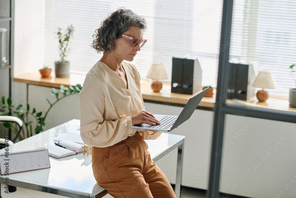 Poster serious mature female analyst in quiet luxury attire typing on laptop keyboard while sitting on desk