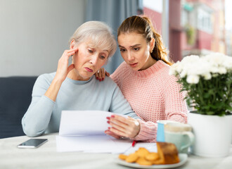 Worried elderly woman sitting in dining room with adult daughter, checking utility bills.