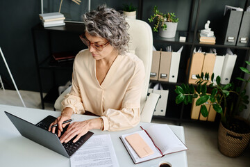 Mature female analyst or broker typing on laptop keyboard while sitting by workplace with financial...