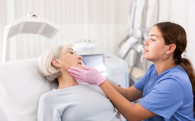 Obraz na płótnie Canvas Positive aged woman patient lying on clinical chair discussing face aesthetic procedures with a specialist