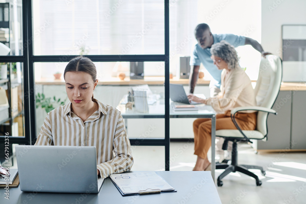 Leinwandbilder Young secretary or analyst using laptop while sitting by desk with financial document with graphic data and looking through information on screen