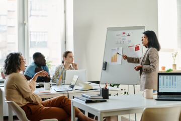Young confident female analyst pointing at paper document with financial chart on whiteboard while explaining data to group of colleagues