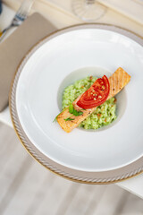 Grilled Salmon Fillet with Green Risotto and Tomato Garnish. Elegant main course featuring grilled salmon fillet served over creamy green risotto and topped with sliced tomato. Presented in a white de