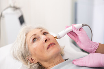 Careful young woman doctor conducting radio frequency lifting procedure on aged woman patient's face