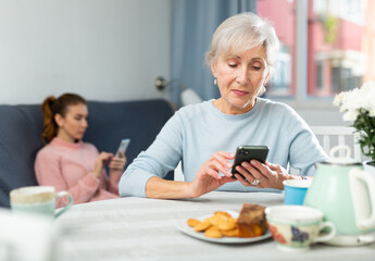 Senior woman sitting at table at home and using her smartphone. Her granddaughter sitting on sofa in background and using smartphone too.
