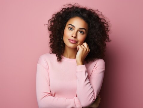 A young woman with voluminous curly hair poses against a pink backdrop. She's wearing a pink top and gazing upwards with a contemplative expression.