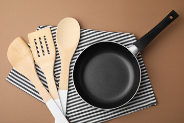 Different kitchen utensils, napkin and frying pan on light brown background, flat lay