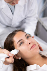 Naklejka premium Delighted young woman lying on clinical chair in aesthetic cabinet during vacuum cleaning of her face