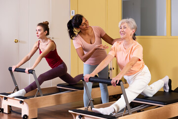 Fototapeta na wymiar Two health conscious women of different ages perform a Pilates exercise using a reformer bed, where a female instructor ..helps them do it correctly