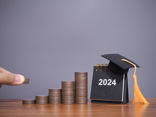Close up hand putting coin in stack of coins, 2024 Desk calendar with graduation hat. The concept of saving money for education, student loan, scholarship, tuition fees in New Year 2024 - obrazy, fototapety, plakaty