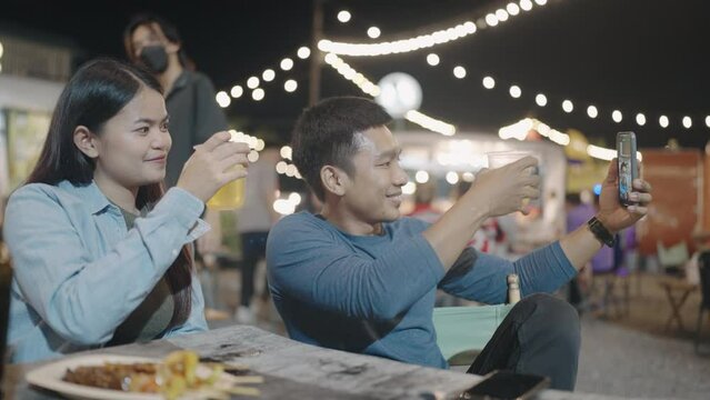 Happy young Asian couple drinking beer alcohol and taking selfie with smartphone in night market