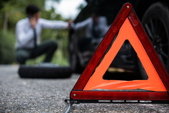 Close-up of a warning triangle in front of a stranded car on the road. Businessman repairing the car engine while standing beside the car. Transportation and roadside assistance concept.