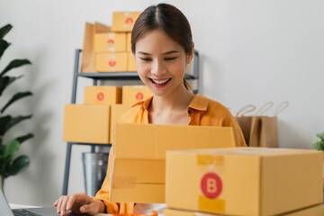 Startup small business, Young Asian woman holding packing boxes and checking online order on digital laptop for products to send to customers. working at the home office.